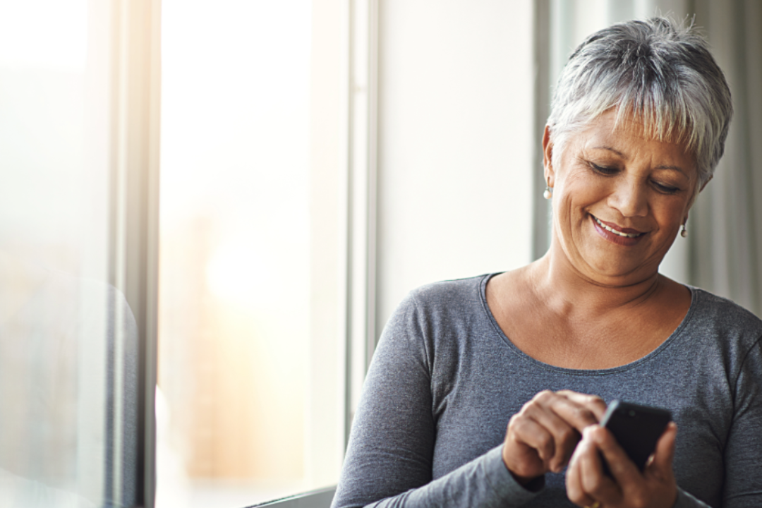 Window, typing and senior woman with a cellphone, social media and mobile app at home. Mature female, happy model and old lady with a smartphone, email and happiness with network and online reading